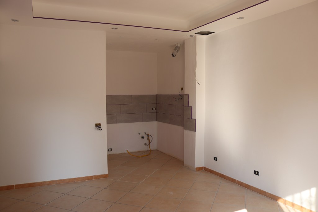 Interno di una casa in fase di ristrutturazione, con pareti bianche e un'area con piastrelle grigie in basso. Esposti tubi e cavi elettrici in evidenza.