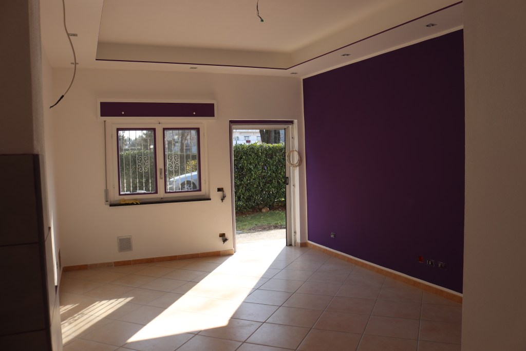 Interno di una stanza con pareti bianche e un muro viola, dotato di una finestra e una porta che si affaccia su un giardino esterno.