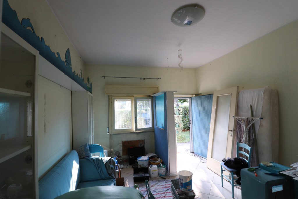 Interno di una casa in fase di ristrutturazione, con divano blu, finestre aperte e vari materiali di lavoro sparsi, inclusi secchi e strumenti.