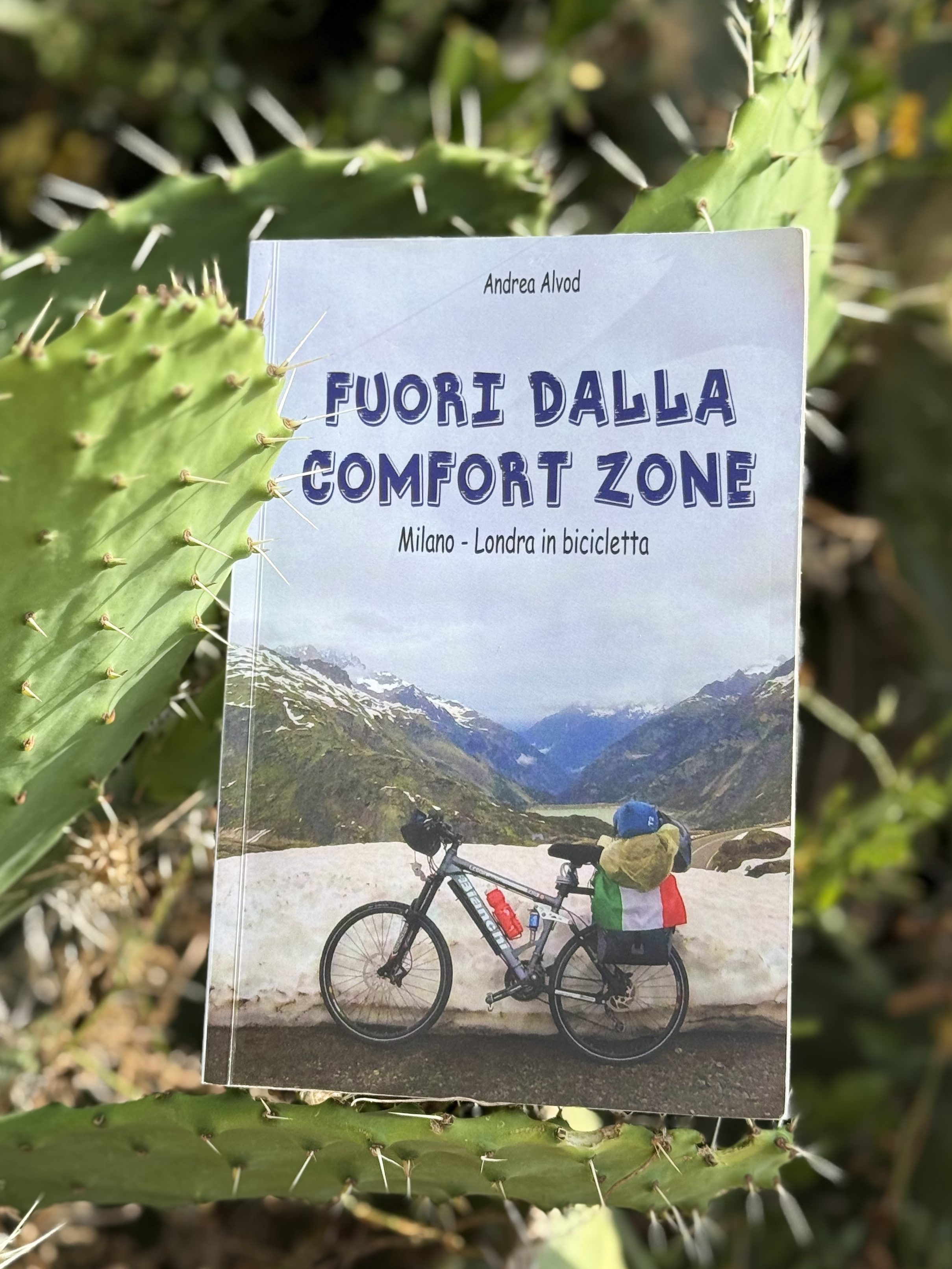 Un libro intitolato 'Fuori dalla comfort zone' di Andrea Alvod, appoggiato su una pianta di cactus, con una bicicletta in primo piano e montagne sullo sfondo.