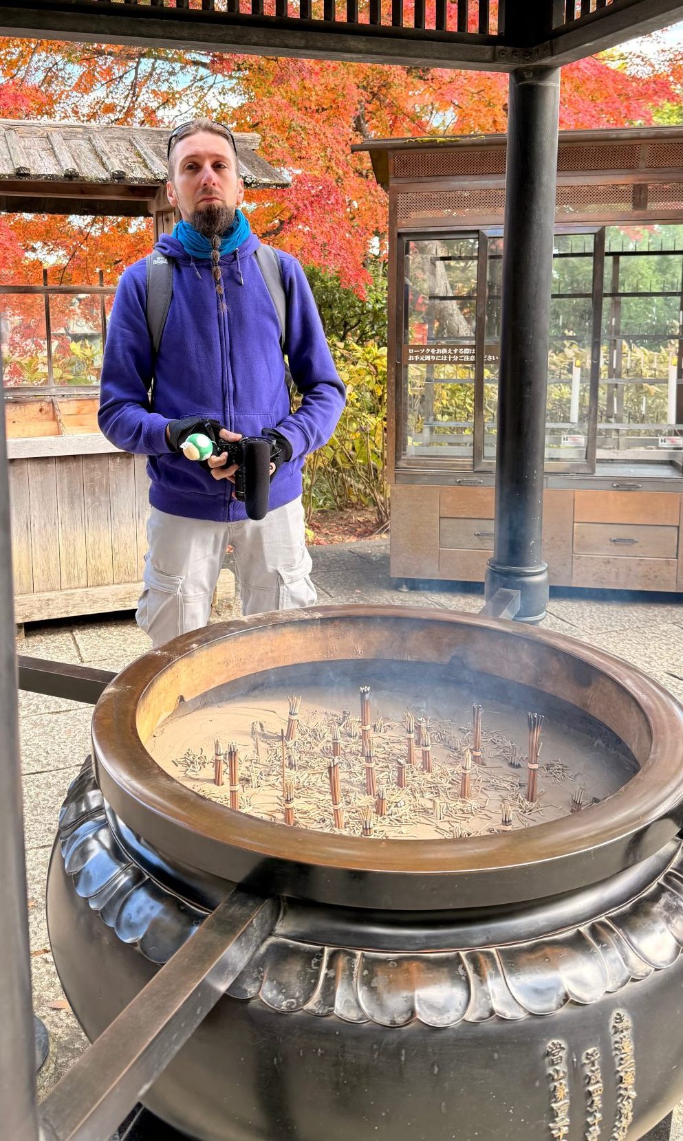 Uomo in un giardino con alberi dai colori autunnali, mentre tiene una fotocamera e si trova accanto a un contenitore di incenso.