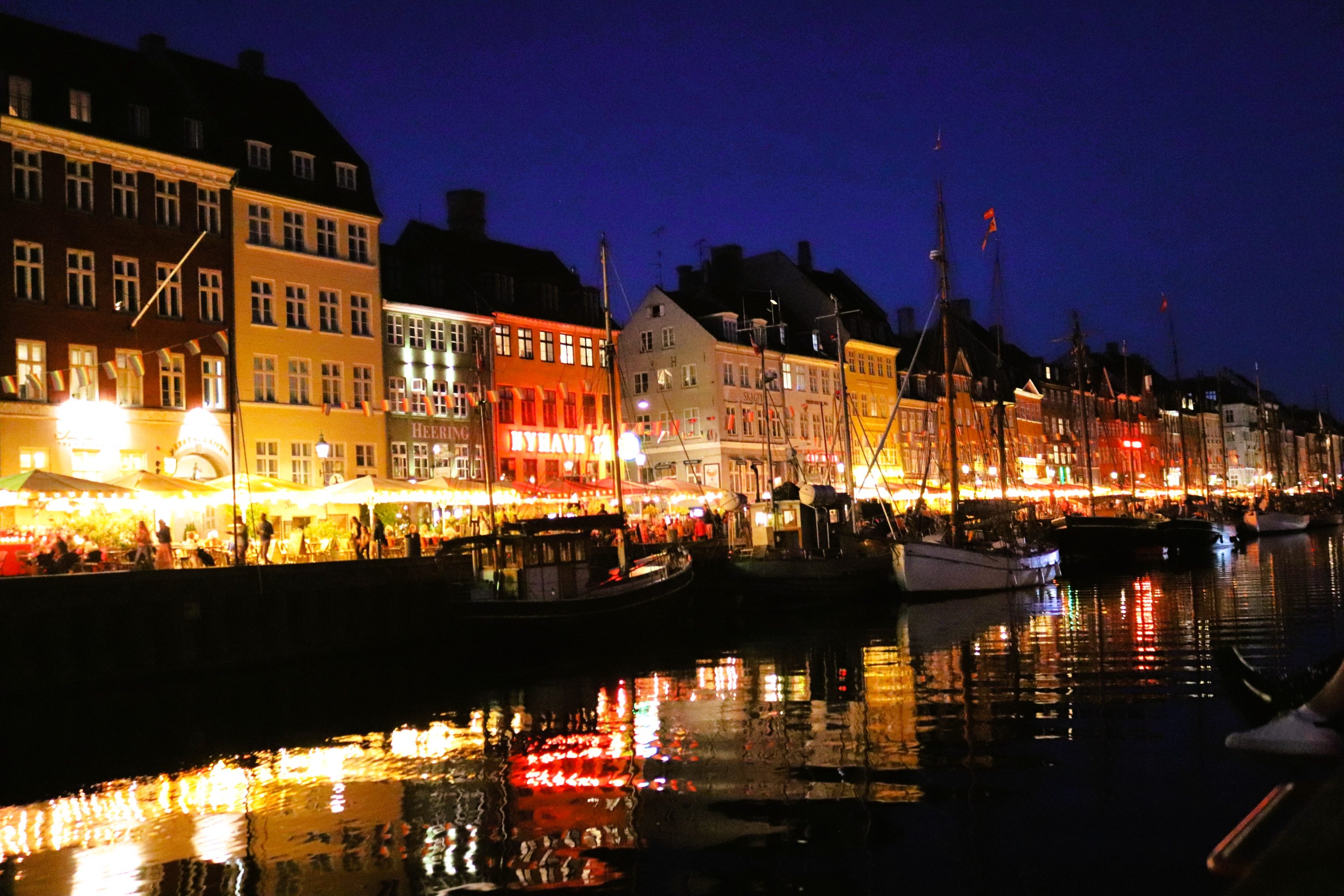 Vista notturna di Nyhavn a Copenaghen, con ristoranti e bar illuminati lungo il canale, e imbarcazioni ormeggiate.