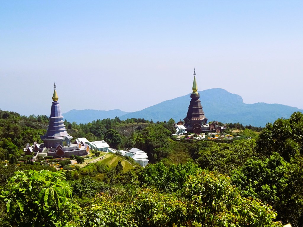 Two pagodas set on a mountain surrounded by lush greenery and distant hills against a clear blue sky.