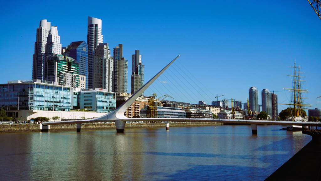 A view of the modern skyline of Buenos Aires featuring tall buildings, with the iconic Puente de la Mujer bridge crossing the water and a tall masted ship docked nearby.