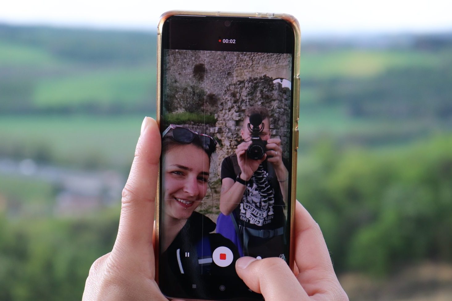 A person holds a smartphone displaying a selfie with a scenic background, capturing another person who is taking a picture with a camera.