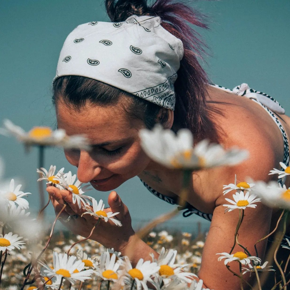 Una persona che indossa una bandana bianca sta annusando dei fiori di margherita in un campo fiorito.