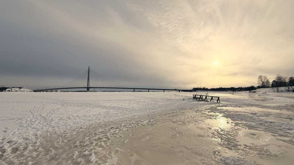 Mare ghiacciato a Helsinki al tramonto, con il ponte strallato all’orizzonte e una scaletta di legno sul ghiaccio